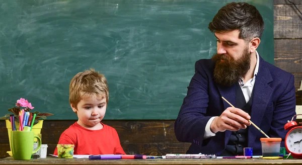 Child and teacher on busy face painting, drawing. Art lesson concept. Teacher with beard, father teaches little son to draw in classroom, chalkboard on background. Talented artist spend time with son