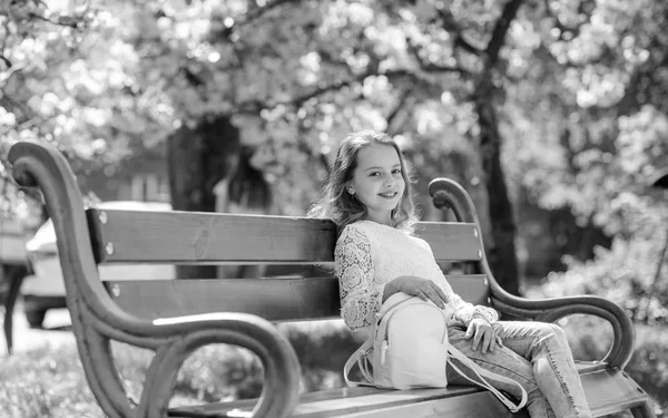 Koncepcja akcesoria. Relaks podczas spaceru w parku, w pobliżu wiśniowy kwiat dziewczyna. Słodkie dziecko z plecaka cieszyć się słoneczny wiosenny dzień. Dziewczyna na uśmiechniętą twarz siedzi na ławce, drzewa sakura na rozmytym tłem — Zdjęcie stockowe