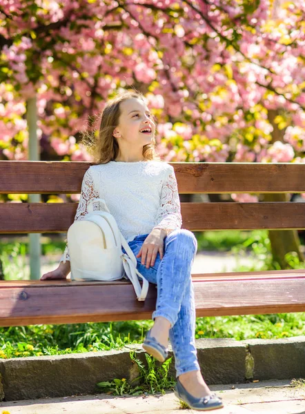 Dziewczyna na uśmiechniętą twarz siedzi na ławce, drzewa sakura na rozmytym tłem. Relaks podczas spaceru w parku, w pobliżu wiśniowy kwiat dziewczyna. Wiosna idzie koncepcja. Słodkie dziecko z plecaka cieszyć się słoneczny wiosenny dzień — Zdjęcie stockowe