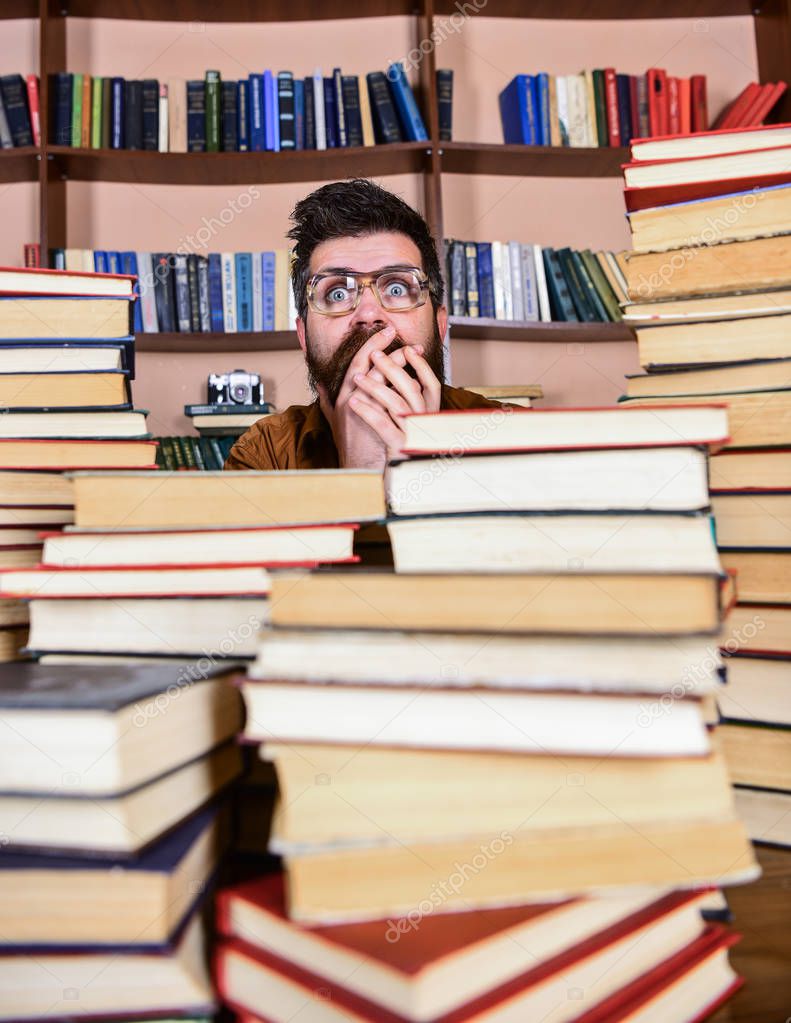 Man on shocked face between piles of books in library, bookshelves on background. Scientific discovery concept. Teacher or student with beard wears eyeglasses, sits at table with books, defocused