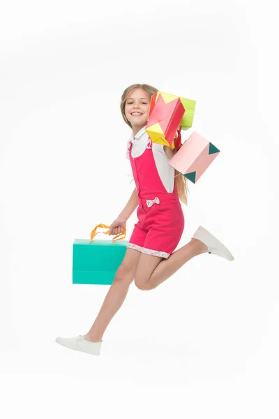 Девушка в розовом комбинезоне прыгает на белом фоне. Улыбающийся ребенок с разноцветными бумажными пакетами. Ребёнок получает подарки на день рождения. Девушка с длинными светлыми волосами, излучающая радость, счастливая детская концепция — стоковое фото
