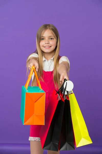 Chica sosteniendo coloridas bolsas de compras. Modelo con pelo largo y rubio sobre fondo violeta. Niño dando regalos en bolsas de papel, el estado de ánimo de vacaciones. Pequeño comprador gran gastador, moda y concepto de ventas — Foto de Stock