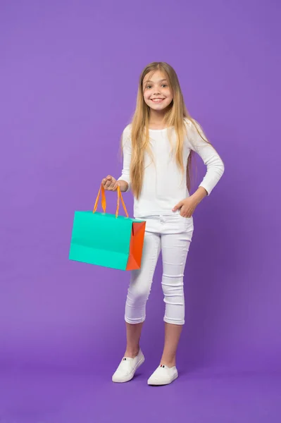 Dívka, která nosí bílý svetr a kalhoty izolované na fialovém pozadí. Dítě s veselou tváří drží papírový sáček. Dítě překvapil dárek, narozeniny koncept. Dívka s velkým úsměvem dělá nakupování — Stock fotografie