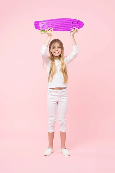 Дівчина тримає фіолетовий скейтборд над головою. Чемпіонка в вуличній гонці, щаслива концепція дитинства. Малюк у білому джемпері, джинси та кросівки ізольовані на рожевому фоні. Дитина з досить усміхненим обличчям — стокове фото