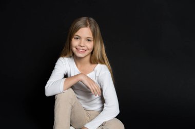 Siyah arka planda moda saç stili olan mutlu çocuk. Küçük kız uzun sarı saçlarıyla gülümsüyor. Güzel çocuk sevimli bir bakışla gülümsüyor. Güzellik salonu. Sakin ol ve saçını yaptır.