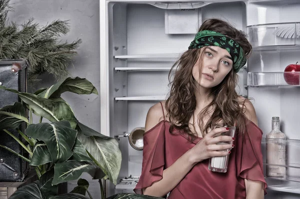 Mulher com aparência cansada de pé ao lado da geladeira aberta. Menina segurando copo de leite. Senhora com cabelo encaracolado e cabeça verde na cozinha, comida e dieta conceito. Grande planta verde na sala de jantar interior — Fotografia de Stock