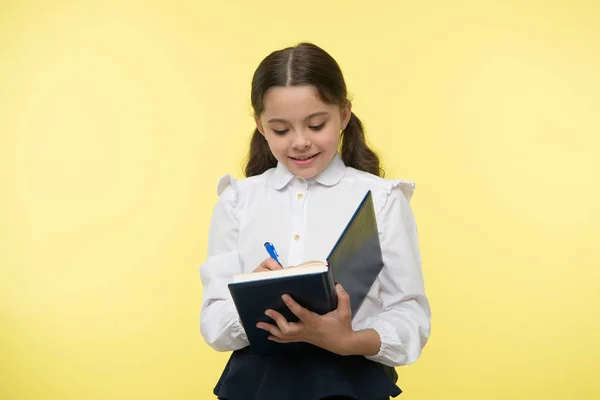 Colegiala escribir en cuaderno sobre fondo amarillo. Chica con uniforme escolar, de vuelta a la escuela. Niño pequeño con almohadilla para escribir. Un genio de la moda. Creando mi futuro —  Fotos de Stock