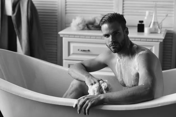 Guy dans la salle de bain avec articles de toilette et chaise avec veste sur fond. Homme avec barbe et visage sérieux prenant un bain. Macho assis nu dans la baignoire comme concept de sexualité et de relaxation — Photo