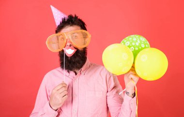 Hava balonları ile parti şapkalı adamı kutluyor. Hippi dev güneş gözlüğü kutluyor. Neşeli yüzündeki sakallı gülümseyen ağız sopa, kırmızı bir arka plan üzerinde tutar. Fotoğraf booth eğlence kavramı