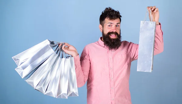 Man with happy face and trendy beard holding shopping bags. Shopaholic spending all salary on clothes. Bearded man in pink shirt isolated on blue background. Man with hipster beard enjoying shopping
