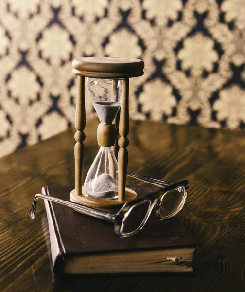 La sabiduría atribuye concepto. Reloj de arena, libro viejo y anteojos en mesa de madera, fondo patrón. Arena cayendo dentro del reloj de arena. Reloj de arena contando el tiempo cerca de anteojos anticuados —  Fotos de Stock