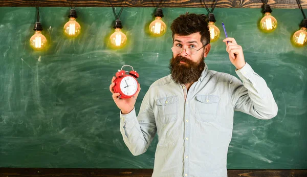 Homem com barba e bigode na cara esquecida comichão na cabeça. O hipster barbudo segura relógio, quadro no fundo, espaço de cópia. Conceito de disciplina. Professor em óculos detém despertador e caneta — Fotografia de Stock