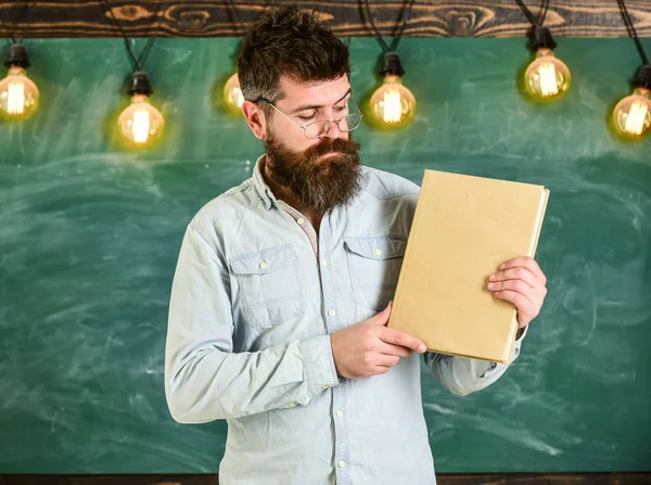 大胡子的时髦人持有书, 黑板上的背景。老师在眼镜上呈现空白的书。文学表现概念。有胡子和胡子的男人在忙碌的脸上站在教室里 — 图库照片