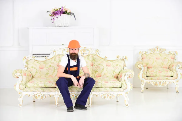 Loader sit on sofa, having rest. Break and rest concept. Man with beard, worker in overalls and helmet sitting on couch tired, white background. Courier relaxing while moving furniture, relocation