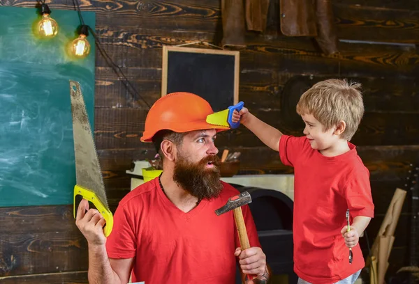 Отец, родители с бородой в защитном шлеме учат маленького сына использовать разные инструменты в школьной мастерской. Ребенок веселый, играет с игрушечной пилой, учится пользоваться инструментами с папой. Концепция отцовства — стоковое фото