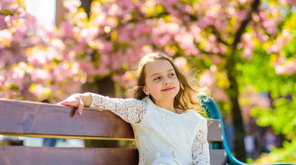 Våren koncept. Flicka på leende ansikte sitter på bänken, sakura träd på bakgrund, oskärpa. Flicka avkopplande medan promenad i park nära körsbärsblommor. Söta barn njuta av soliga vårdag — Stockfoto