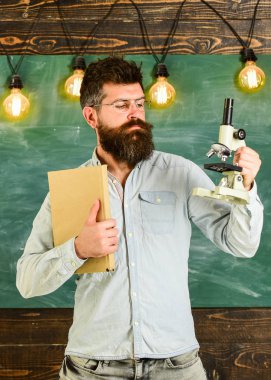 Gözlük öğretmen kitap ve mikroskop tutar. Sakal ve bıyık sınıfta sıkı yüzünde olan adam. Bilim adamı kitap ve mikroskop, kara tahta arka plan üzerinde tutar. Bilimsel araştırma kavramı