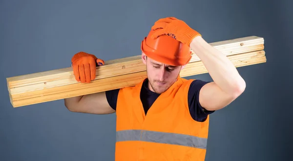 Safety and protection concept. Man in helmet, hard hat and protective gloves holds wooden beam, grey background. Carpenter, woodworker, strong builder on serious face carries wooden beam on shoulder