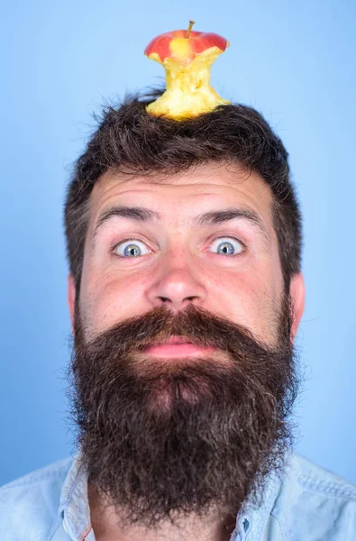 Hipster happy face with apple stump target on head blue background, close up. Man handsome hipster long beard almost eaten apple stump on head as target. Weight loss goal. Live target concept