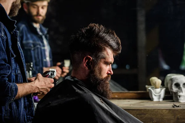 Barber with clipper styling hair of brutal bearded client. Hipster lifestyle concept. Barber with hair clipper works on hairstyle for bearded man barbershop background. Hipster client getting haircut