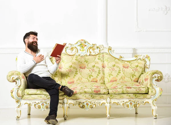 Un tipo leyendo un libro viejo con placer. Hombre con barba y bigote se sienta en el sofá de estilo barroco, sostiene libro, fondo blanco de la pared. Macho en el libro de lectura de caras de risa. Concepto de literatura humorística —  Fotos de Stock