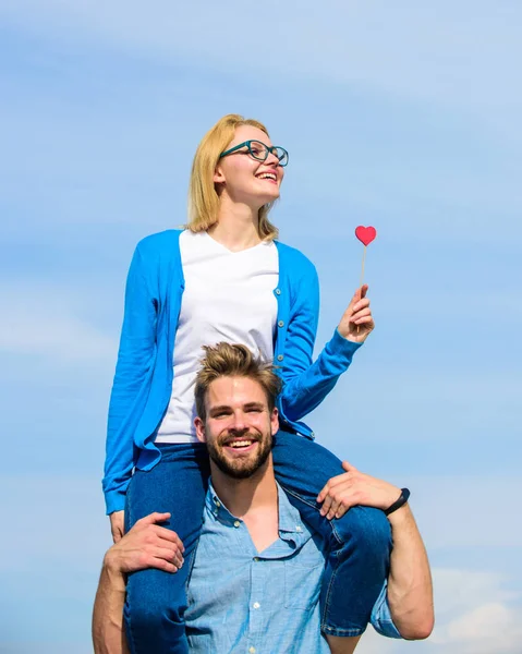 L'uomo porta la ragazza su spalle, sfondo di cielo. Coppia felice appuntamento divertirsi insieme. Donna tiene il cuore sul bastone simbolo di amore. Concetto di appuntamento romantico. Coppia innamorata a piedi giornata di sole all'aperto — Foto Stock