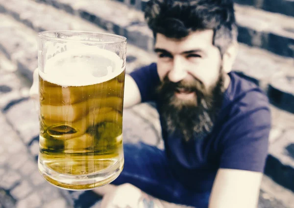 Um tipo a levantar copos com cerveja. Hipster no rosto sorridente bebe cerveja ao ar livre. Um conceito de saúde. Homem com barba e bigode segura vidro com cerveja enquanto se senta em escadas de pedra, desfocado — Fotografia de Stock