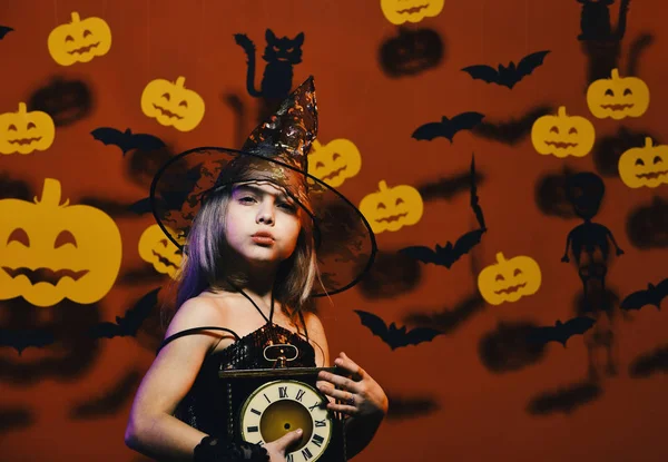 El chico con disfraz de bruja espeluznante sostiene el reloj viejo. Fiesta de Halloween y decoración concepto. Pequeña bruja con sombrero negro. Chica con la cara preocupada — Foto de Stock