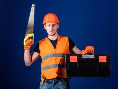 Kask, kask içinde adam araç kutusu taşır ve el testeresi, mavi arka plan tutar. Marangoz kavramı. İşçi, repairer, tamirci üzerinde ciddi bir yüz taşıyan araç, onarım için kopya alanı hazır