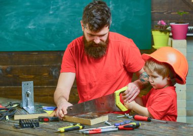 Masculine duties concept. Father, parent with beard teaching little son to sawing with sharp handsaw, carpenter crafts. Boy, child busy in protective helmet learning to use handsaw with dad clipart