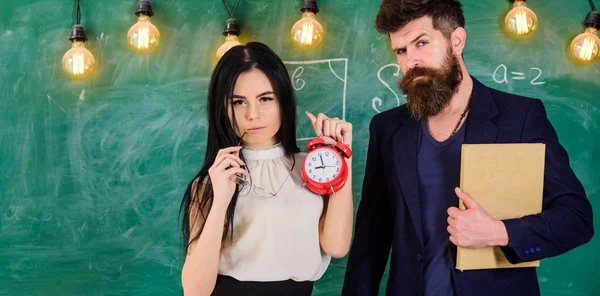 Man with beard hold book and sexy girl teacher holds alarm clock, chalkboard on background. School rules concept. Lady teacher and strict schoolmaster care about discipline and rules in school — Stock Photo, Image