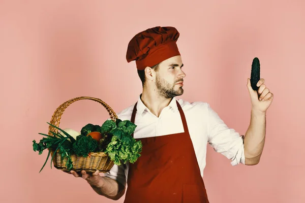Kock med nyfikna ansikte innehar gurka och flätade korg med färska grönsaker. Kock i Bourgogne enhetliga ser på gurka i hand. — Stockfoto