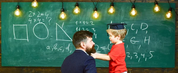 Young man and small boy side view in the classroom. Kid pulling beard of the guy. Cute child in graduation cap in front of the green board — Stock Photo, Image