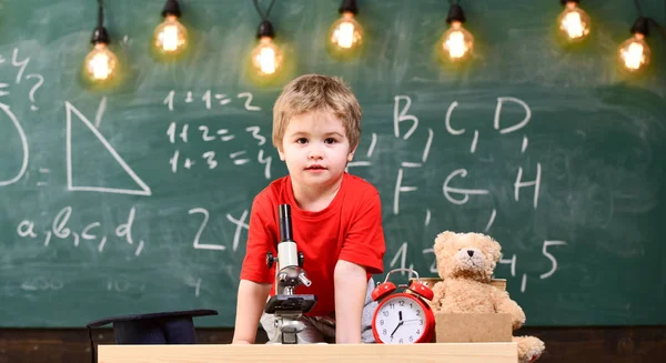 Första tidigare intresserade studera, lärande, utbildning. Barn på glada ansikte nära klocka och Nalle. Grundskola-konceptet. Kid boy nära mikroskopet i klassrummet, svarta tavlan på bakgrund — Stockfoto