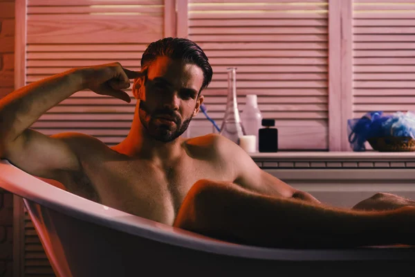Typ im Badezimmer mit Toilettenartikeln im Hintergrund, selektiver Fokus. Sex- und Erotikkonzept: Macho sitzt nackt in Badewanne — Stockfoto