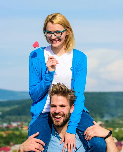 El hombre lleva a su novia en hombros, fondo del cielo. Concepto de fecha romántica. La mujer sostiene el corazón en el palo símbolo del amor. Pareja feliz cita divirtiéndose juntos. Pareja enamorada caminando al aire libre día soleado —  Fotos de Stock