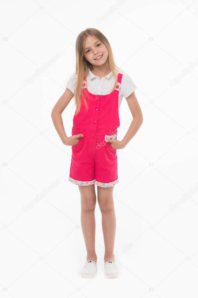 Girl wearing hot pink jumpsuit, white blouse and shoes. Cheerful kid in summer outfit, vacation concept. Child holding hands in pockets. Lovely girl with long blond hair isolated on white background