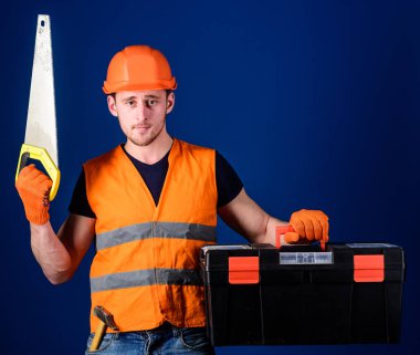 Kask, kask içinde adam araç kutusu taşır ve el testeresi, mavi arka plan tutar. İşçi, repairer, tamirci üzerinde ciddi bir yüz taşıyan araç, onarım, kopya alanı için hazır. Marangoz kavramı