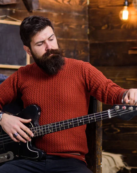 Músico de rock com aparência brutal posando com instrumento. Homem barbudo afinando guitarra elétrica. Homem com barba elegante e bigode em camisola de malha terracota e calça jeans preta relaxante, conceito de música — Fotografia de Stock