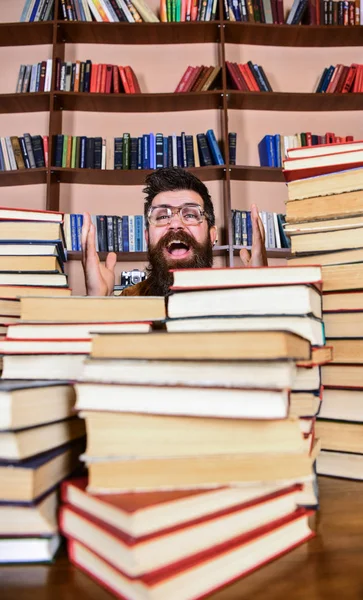 Pojęcia naukowe odkrycia. Człowiek na twarzy podekscytowany między stosy książek w bibliotece, regały na tle. Nauczyciel lub student z brodą nosi okulary, siedzi przy stole z książek, rozmytym — Zdjęcie stockowe
