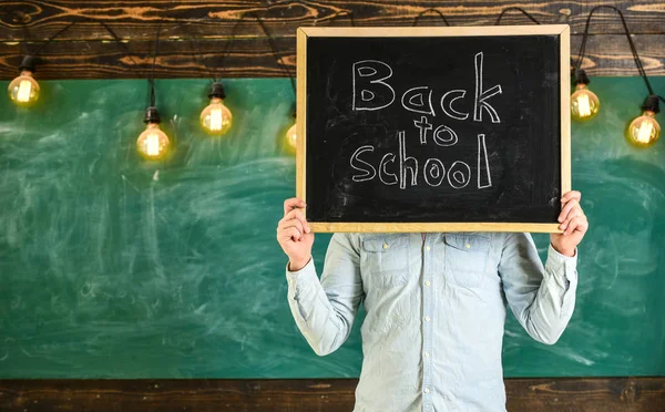 Start of school year concept. Teacher holds chalkboard in front of face. Man welcomes students, chalkboard on background. Teacher faceless holds blackboard with title back to school