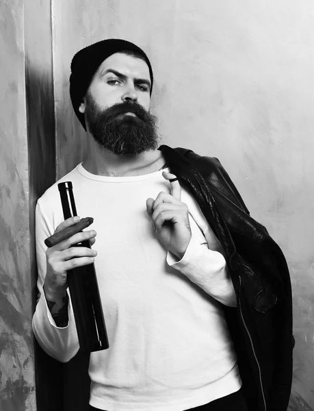 Бородатый брутальный кавказский хипстер держит бутылку и курит сигару. — стоковое фото