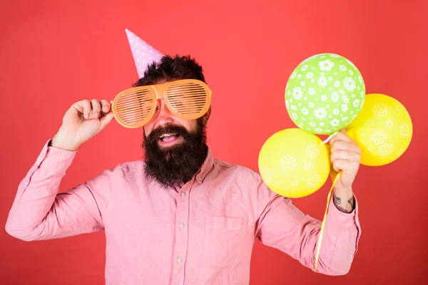 Hipster gluren uit gigantische zonnebril. Man met baard en snor op blij gezicht houdt stelletje lucht ballonnen, rode achtergrond. Viering concept. Guy in feest hoed met lucht ballonnen viert — Stockfoto