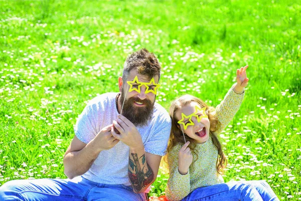 Rock star concept. Father and daughter sits on grass at grassplot, green background. Child and dad posing with star shaped eyeglases photo booth attribute at meadow. Family spend leisure outdoors