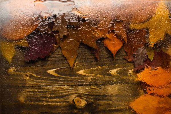 Fondo de otoño, vista a través de vidrio húmedo sobre madera y hojas caídas. Gotas de agua o lluvia sobre vidrio transparente y hojas naranjas de otoño sobre textura de madera. Concepto textura de fondo — Foto de Stock