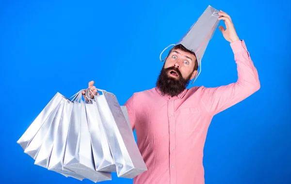 Uomo con barba e baffi porta borse della spesa, sfondo blu. Guy shopping sulla stagione delle vendite con sconti. Concetto di shopping. Hipster sul viso sorpreso con borsa sulla testa è dipendente shopaholic — Foto Stock