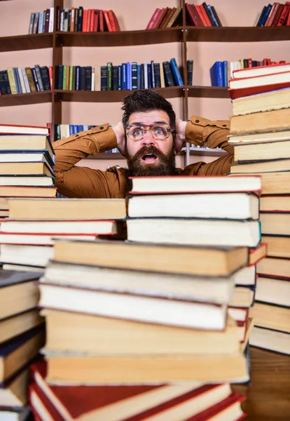 Professor ou estudante com barba usa óculos, senta-se à mesa com livros, desfocado. Homem no rosto chocado entre pilhas de livros na biblioteca, estantes de livros no fundo. Conceito de descoberta científica — Fotografia de Stock