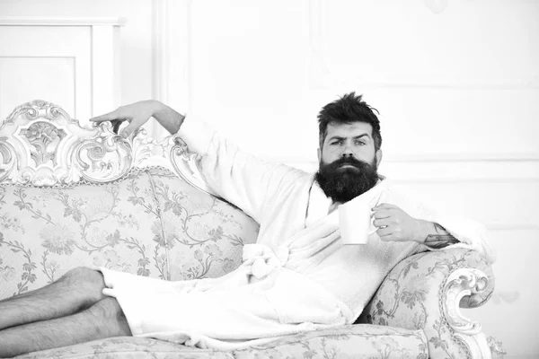 Man with beard and mustache enjoy morning while sitting on old fashioned luxury sofa. Man on thoughtful face in bathrobe drink coffee in luxury hotel in morning, white background. Luxury life concept