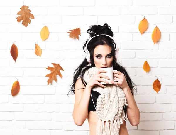 Girl with serious face holds and drinks cup of coffee on white brick wall background. Autumn club party with dry leaves decor. Hot drink and club concept. Dj lady wears black bra and white headphones