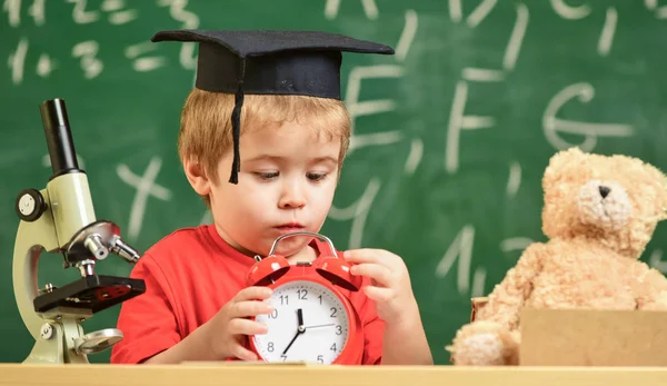 Alumno esperando el receso escolar. Niño en cara triste mira el despertador. Niño en gorra académica cerca del microscopio, sostiene el reloj en el aula, pizarra en el fondo. Concepto de descanso escolar — Foto de Stock